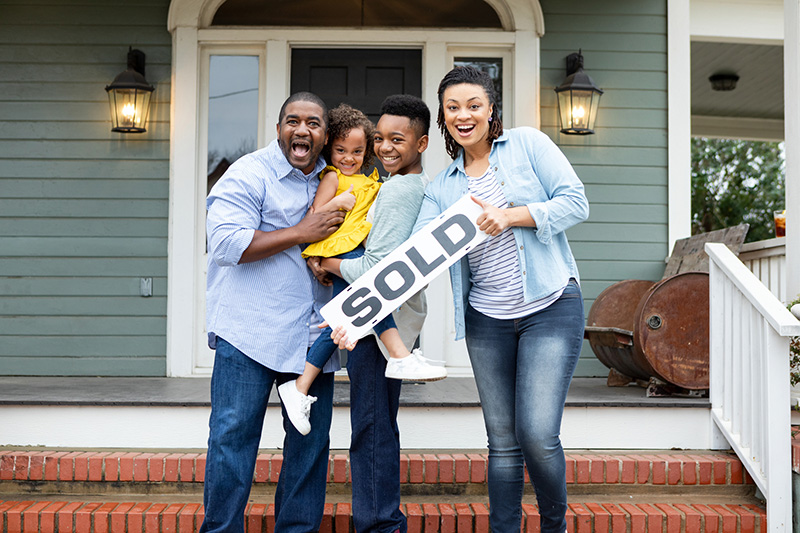 A happy multiracial family stands on the porch of their newly sold home, holding a "SOLD" sign and beaming.