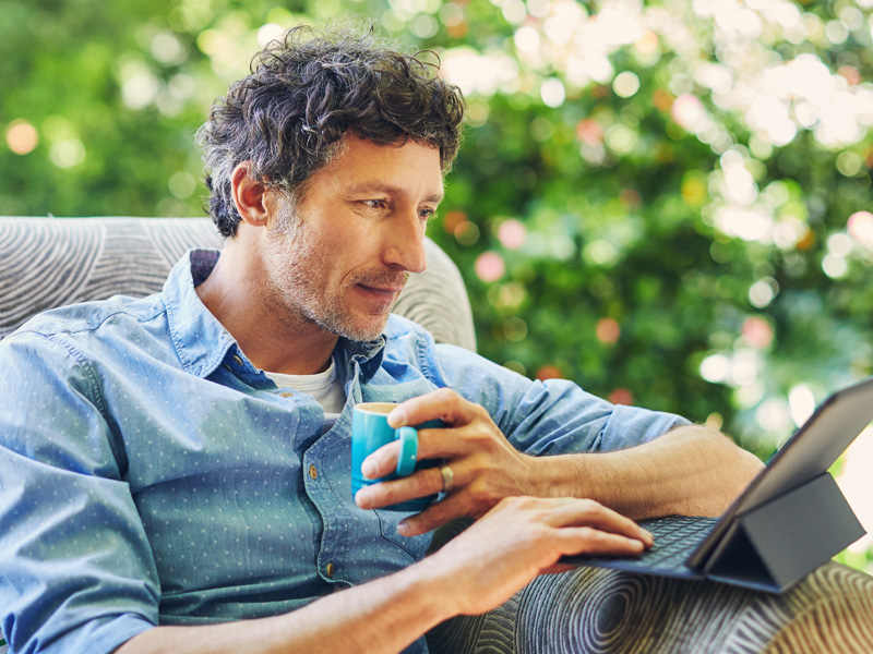 a man sitting outside on a chair with a cup of coffee looking at a device on what is happening in the stock market.